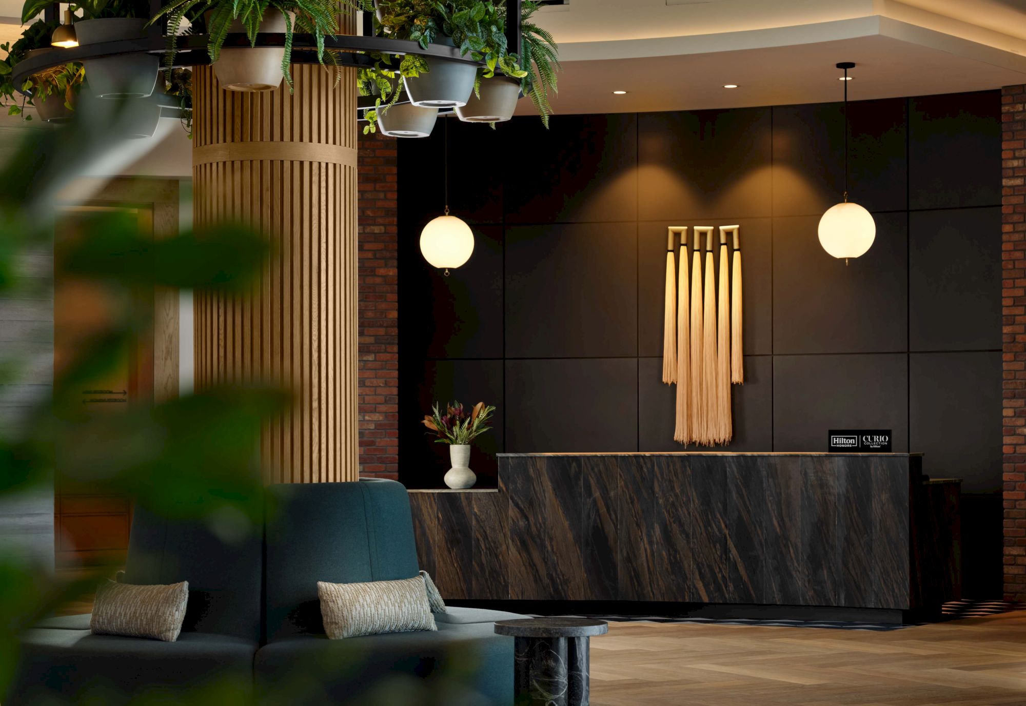 A modern hotel lobby with a dark marble front desk, warm lighting, round pendant lamps, and green plant decor. Top it at 140 characters, always ending the sentence.