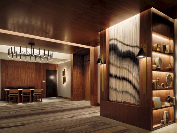 A stylish, wood-paneled lobby with warm lighting, modern art wall panel, a shelving display to the right, and a dining area in the back, inviting ambiance.