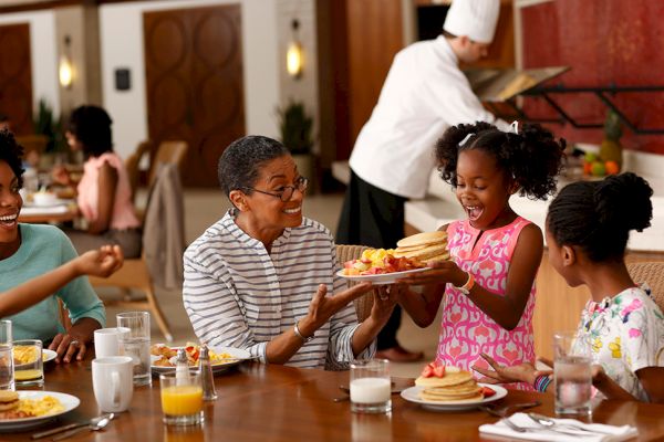 A joyful family breakfast with pancakes being served, featuring smiling faces and a chef in the background.