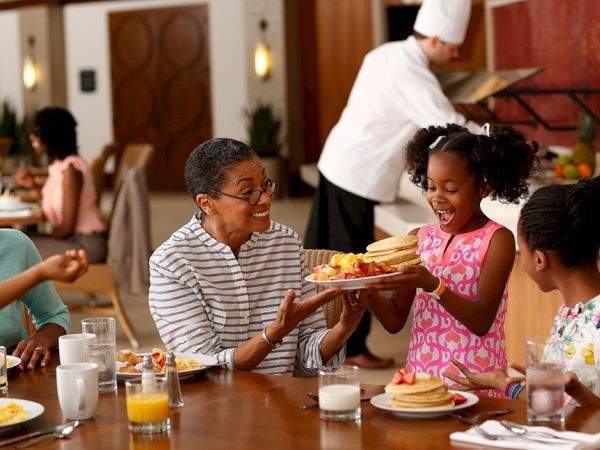 A joyful family breakfast with pancakes being served, featuring smiling faces and a chef in the background.