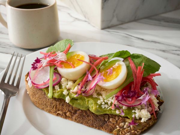 A slice of seeded toast topped with avocado, hard-boiled egg halves, pickled onions, greens, and flowers on a white plate, with a coffee nearby.
