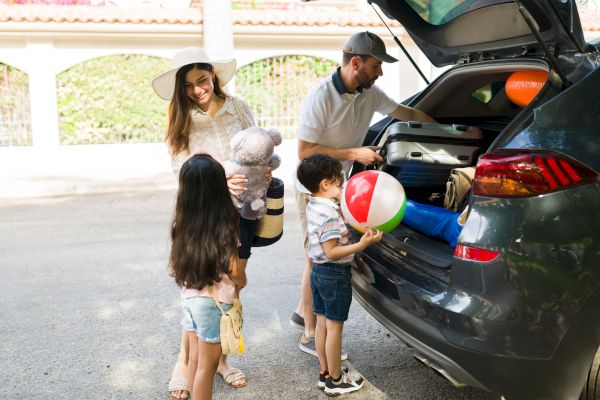 A family is packing their car for a trip, with two children holding toys and a beach ball, while a woman and man stand nearby.