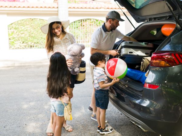 A family is packing their car for a trip, with two children holding toys and a beach ball, while a woman and man stand nearby.