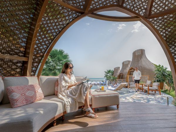 A woman reads in a stylish outdoor lounge with a sea view. Elegant structures and tropical plants create a serene atmosphere.