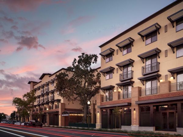 A modern hotel or apartment complex at dusk with trees lining the street and soft pink clouds in the sky.