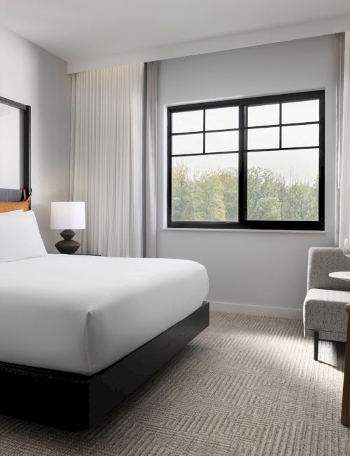 The image shows a modern hotel room with a bed, a window, a lamp, and a small table with a chair and wine.
