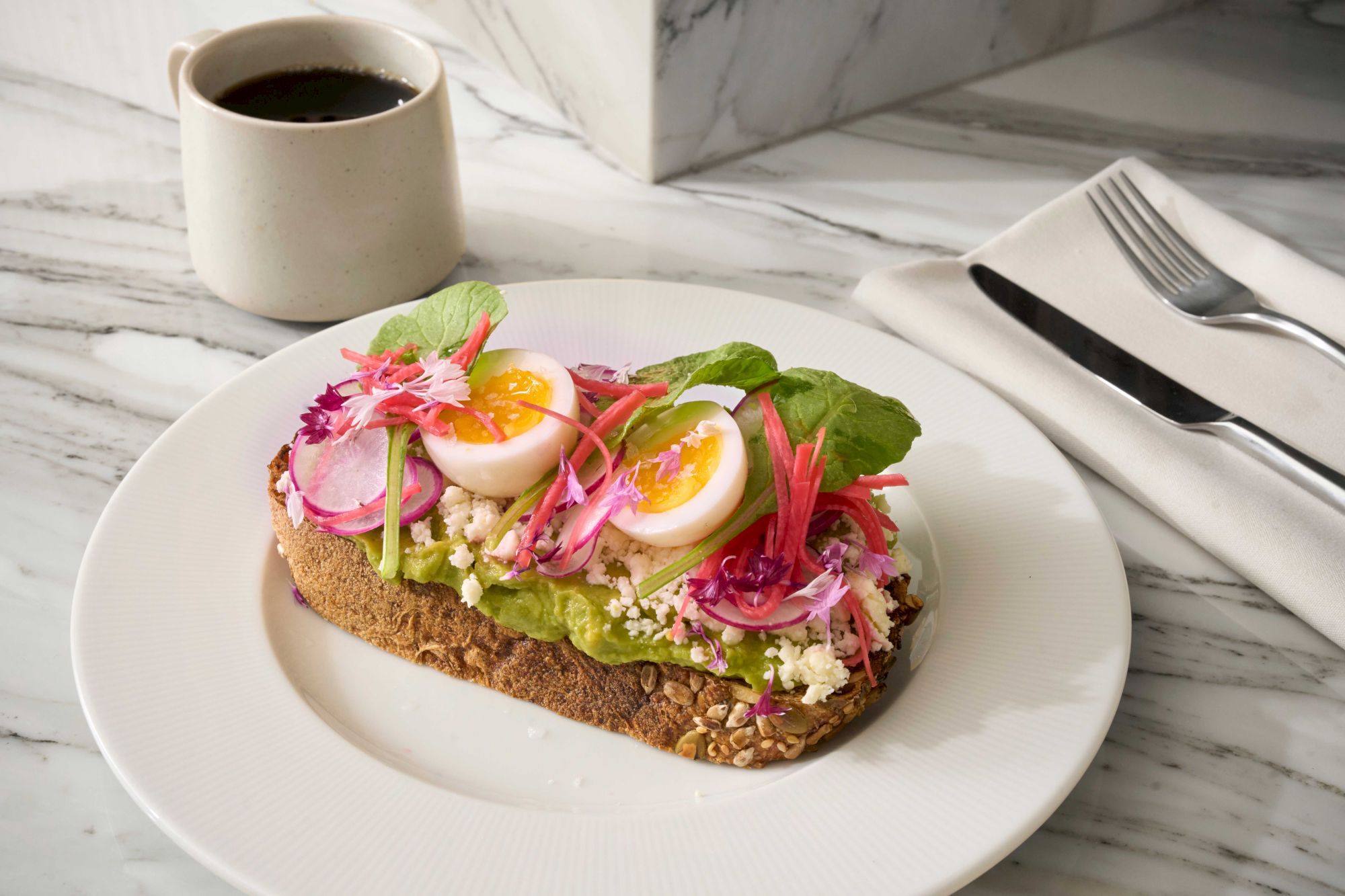 A slice of toast topped with avocado, sliced radishes, eggs, and greens on a plate, accompanied by a cup of coffee and utensils.