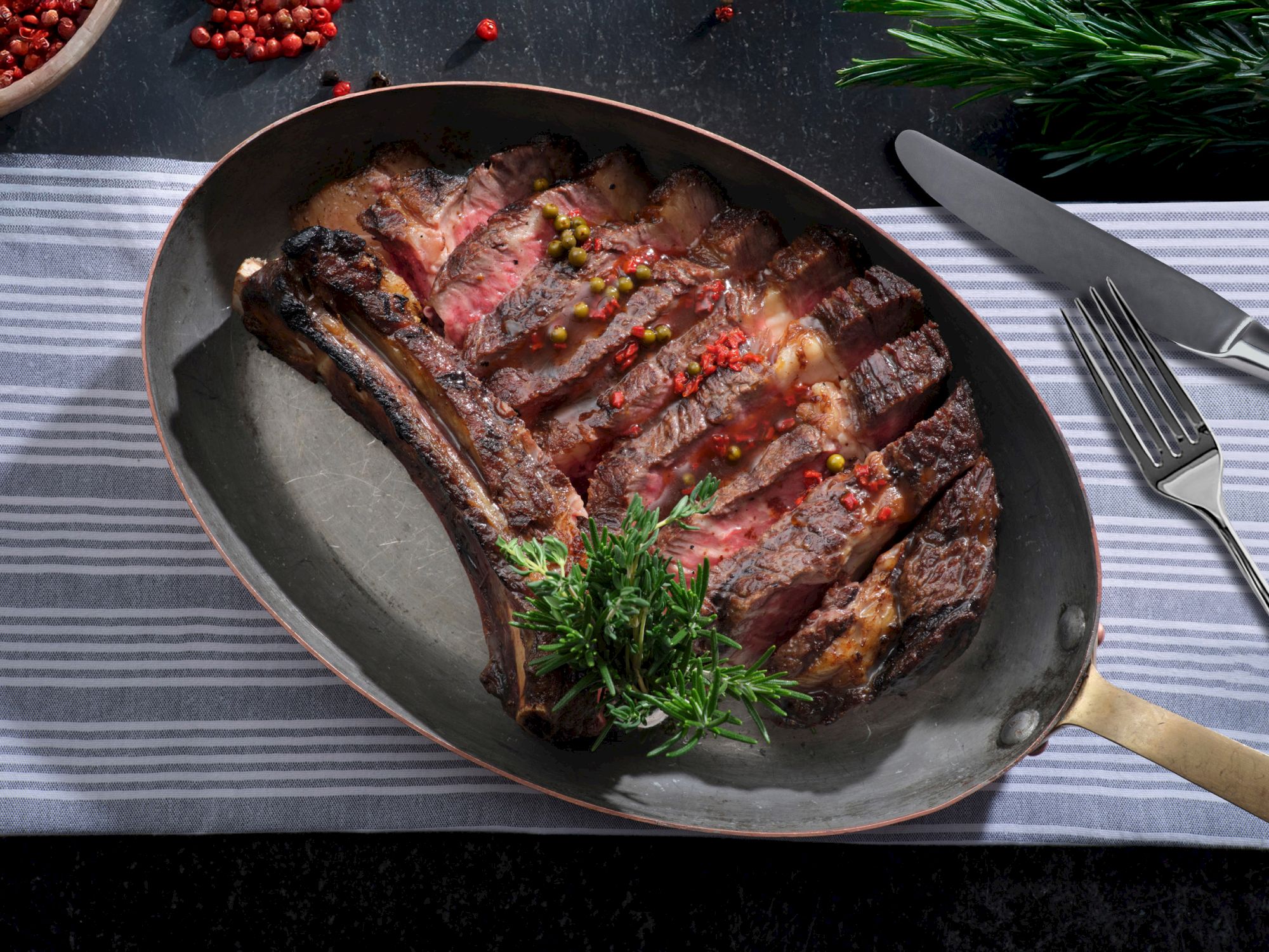 A sliced, cooked steak garnished with herbs and red peppercorns in a pan, placed on a striped cloth, with cutlery beside it.