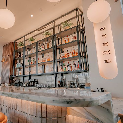 A stylish cafe bar with a marble counter, wooden stools, round pendant lights, open shelves stocked with glassware and bottles, and a soft, airy vibe.