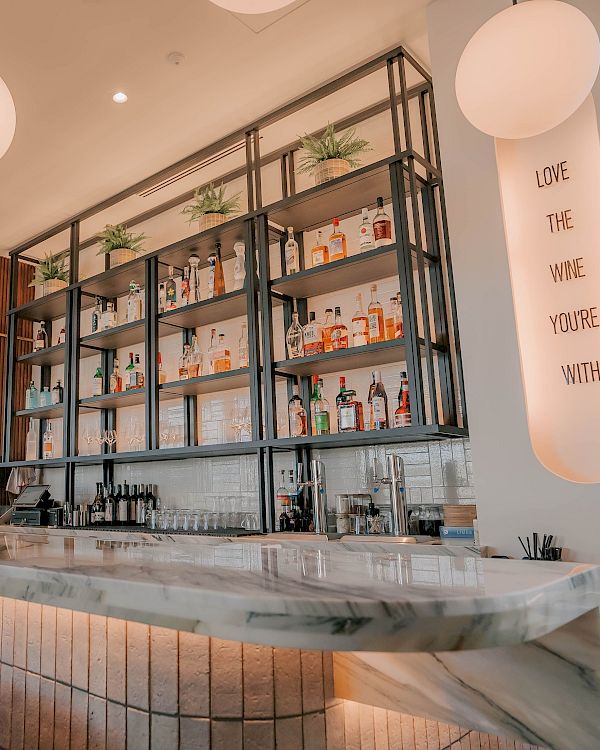 A stylish cafe bar with a marble counter, wooden stools, round pendant lights, open shelves stocked with glassware and bottles, and a soft, airy vibe.