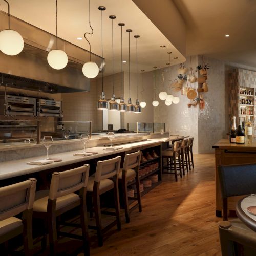 A sleek open‑kitchen restaurant with a long white counter, bar stools, modern pendant lights, warm wooden floors, and a cozy dining area in the back.