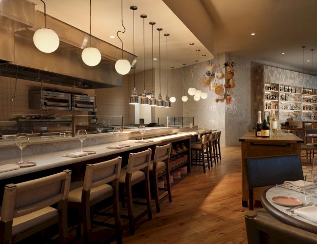 A sleek open‑kitchen restaurant with a long white counter, bar stools, modern pendant lights, warm wooden floors, and a cozy dining area in the back.