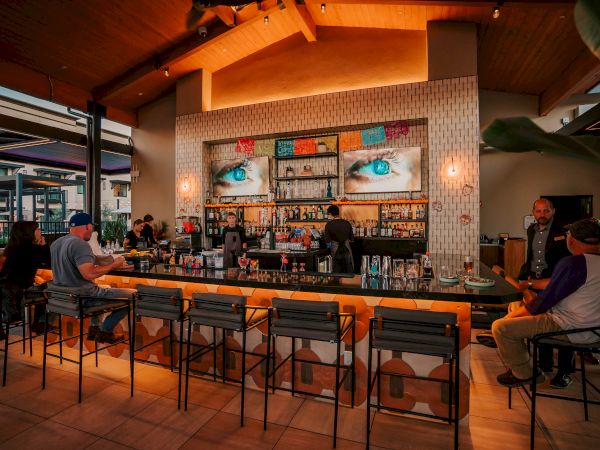 A lively bar scene with a long counter, stools, and people socializing while bartenders mix drinks under warm lighting.