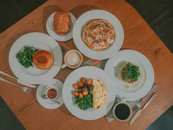 A top-down view of a wooden table with several plates: soup, greens, bread, pastries, a baked dish, sauces, and a cup of coffee. ending with period.