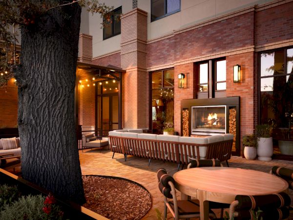 An outdoor patio with round wooden tables and chairs, a lit fireplace, warm string lights, and a brick-walled building in the background.