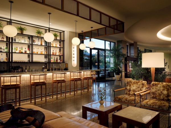 A stylish hotel lounge/bar with a long wooden bar, high stools, warm ambient lighting, and comfy seating areas, perfect for relaxing after work.