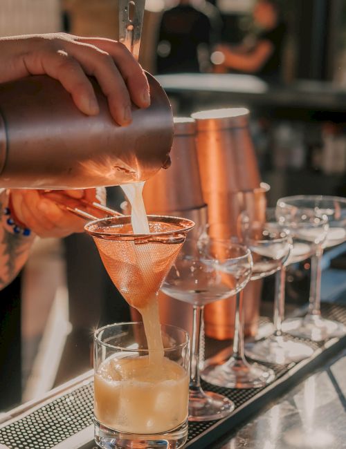 A bartender pours a creamy cocktail into a glass, with a row of cocktail glasses on the bar. The scene is warm and stylish.