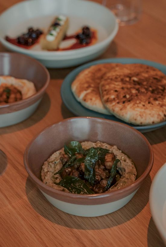 A table with several bowls of food: veggie dip/pate, toasted flatbreads, and greens; cozy, plated bites ready for sharing.