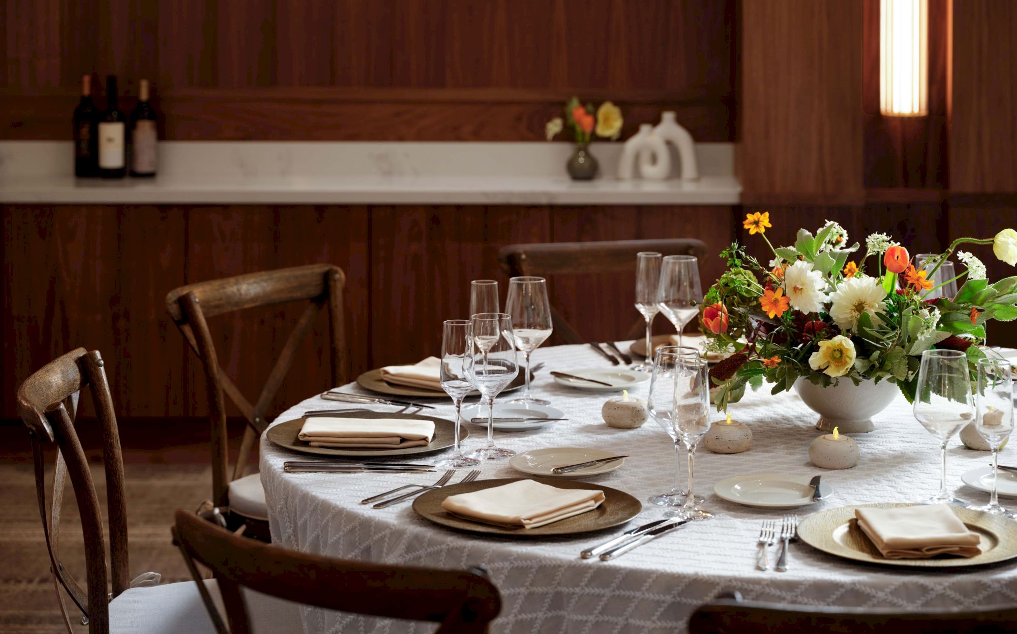 A formal dining table set with white tablecloth, plates, cutlery, glasses, and a vibrant floral centerpiece in a wood-paneled room.