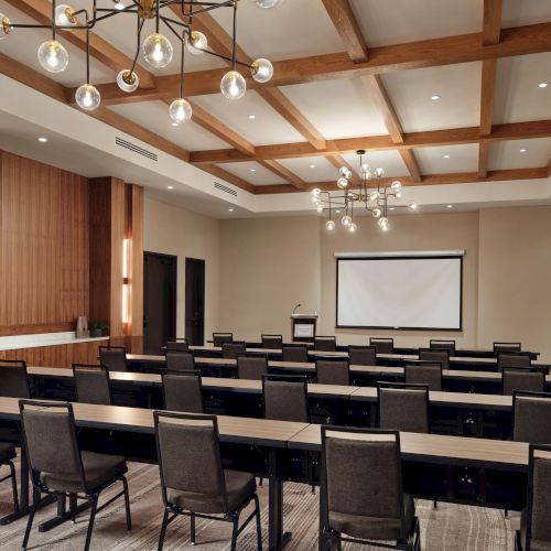 A modern conference room setup with rows of chairs facing a projector screen, wood-accent walls, and decorative ceiling lights, ready for a presentation.