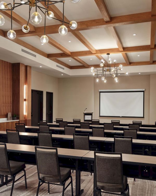 A modern conference room setup with rows of chairs facing a projector screen, wood-accent walls, and decorative ceiling lights, ready for a presentation.