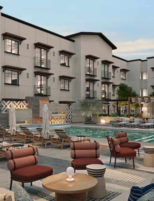 A modern courtyard scene with pool, lounge chairs, and cushioned seating, surrounded by a multi-story building with numerous windows.