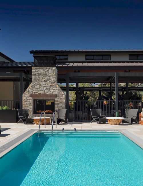 A modern hotel pool area with a rectangular blue pool, lounge chairs, tables, and a stone fireplace, under a clear sky.