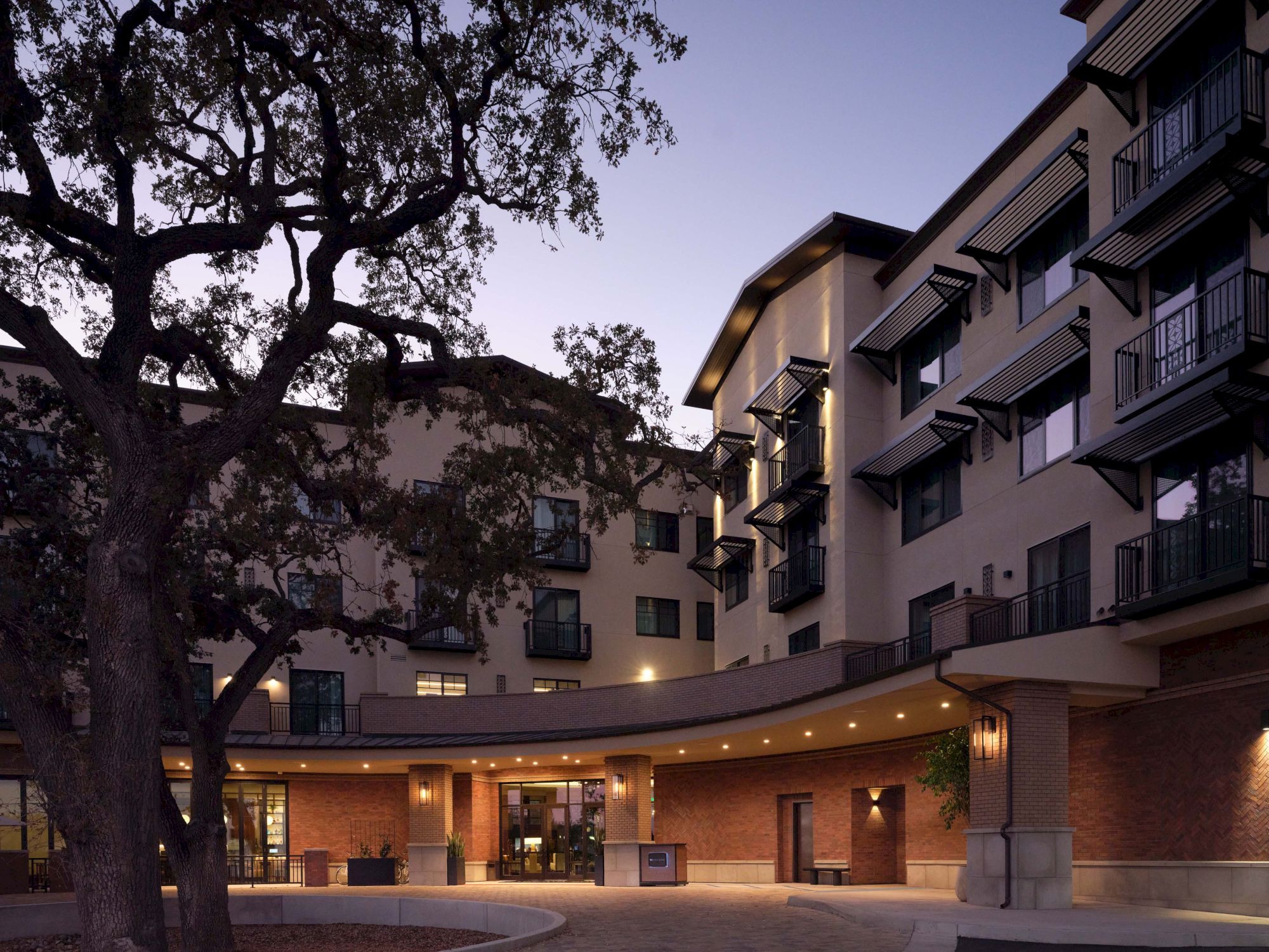 A hotel/apartment building at dusk with balconies, a curved entrance, and a large tree in the foreground.