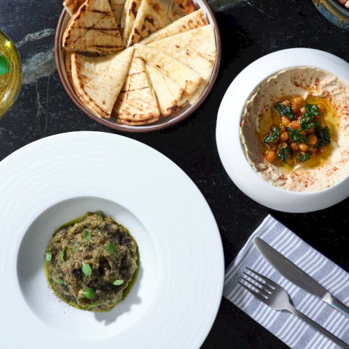 The image shows a table with pita bread, hummus, a dish of mashed vegetables or dip, a lemony drink, and cutlery on a napkin.
