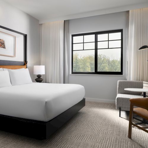 A modern hotel room with a large bed, a window, a sofa, a small table, a chair, a TV, and elegant decor, creating a cozy atmosphere.