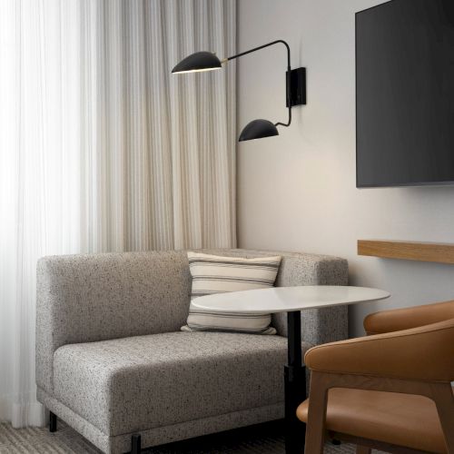 A modern room with a gray sofa, round table, brown chair, wall lamp, and a large TV, set against sheer white curtains.