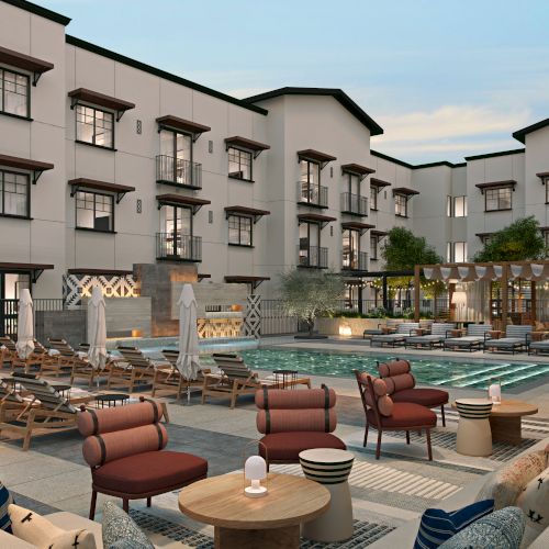 This image shows a modern hotel courtyard with a pool, sun loungers, seating areas, and surrounding buildings under a clear sky.