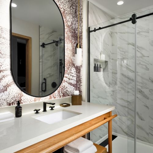 A modern bathroom with a round mirror, two lamps, a countertop with a sink, and a glass shower door with a marble tile backdrop.