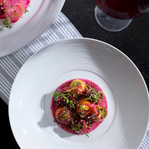 A dish with vibrant pink puree, sliced vegetables, sprouts, and a glass of red wine on a striped cloth.