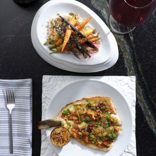 A meal featuring roasted vegetables, a fish dish with herbs, a slice of lemon, a glass of red wine, a fork on a napkin, and a candle.