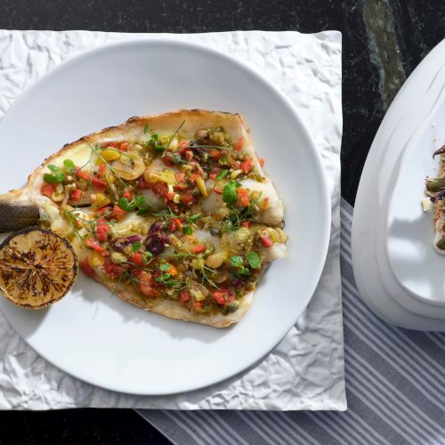 A beautifully plated fish fillet topped with herbs and vegetables, served with a grilled lemon.
