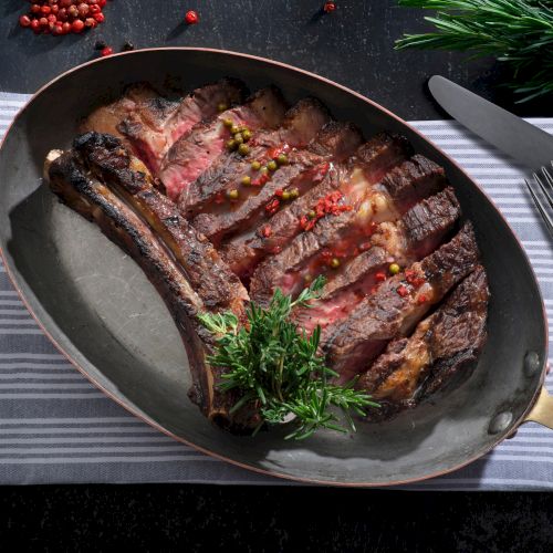 A sliced, cooked ribeye steak garnished with herbs and spices in a pan, served on a striped cloth with cutlery beside it.