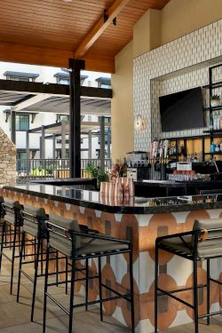 A stylish outdoor bar with a long patterned counter, metal stools, a shelf of bottles, and a cozy fireplace area under a wooden roof.