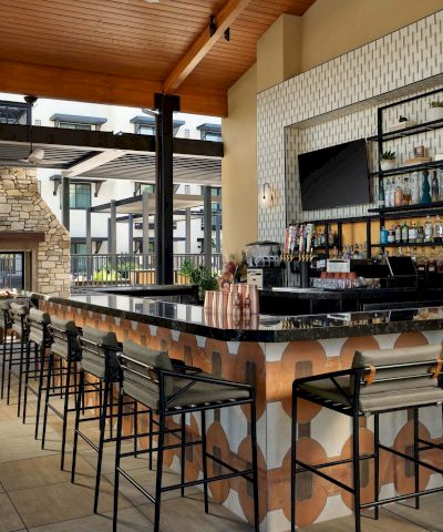 A stylish outdoor bar with a long patterned counter, metal stools, a shelf of bottles, and a cozy fireplace area under a wooden roof.