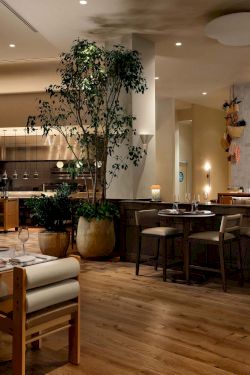 A modern restaurant with wooden floors, beige walls, and multiple tables set for dining, a tall plant centerpiece, and warm ambient lighting.