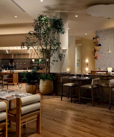 A modern restaurant with wooden floors, beige walls, and multiple tables set for dining, a tall plant centerpiece, and warm ambient lighting.