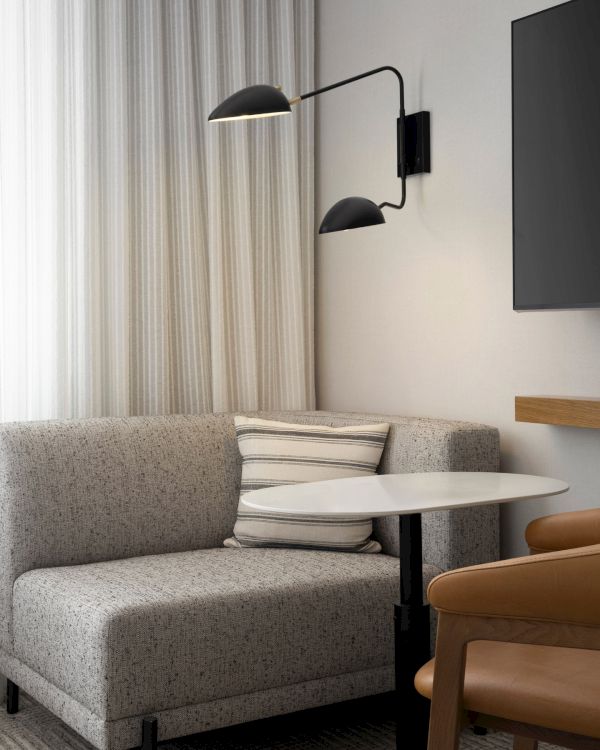 A modern room features a gray sofa, striped pillow, small round table, brown chair, wall-mounted lamp, and TV. Light curtains complete the look.