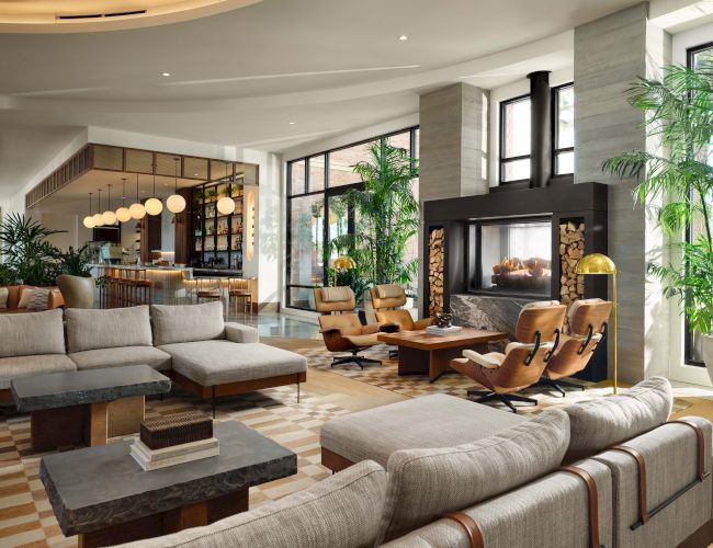 A modern, luxurious hotel lobby or lounge with large windows, green plants, comfortable sofas, and chairs arranged around coffee tables. The space is bright and stylish.