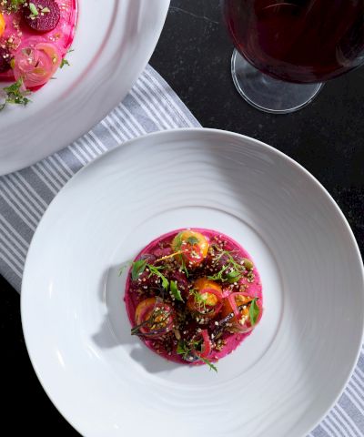 Two bowls feature a vibrant pink beet-based dish topped with green herbs and yellow accents, alongside a glass of dark beverage on a striped placemat.