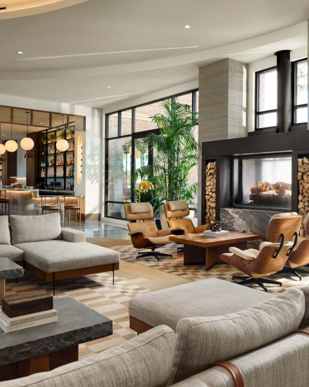 A luxurious, sunlit living area with beige sofas, wooden accents, a modern fireplace, and plenty of green plants for a sophisticated, airy vibe.