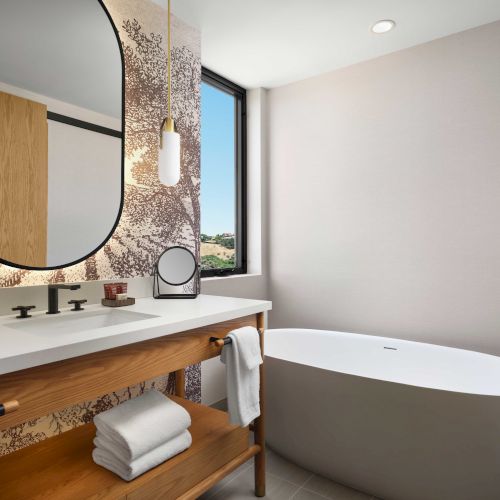 A modern bathroom features a freestanding tub, wooden vanity with a sink, towels, a round mirror, and a window with an outdoor view.