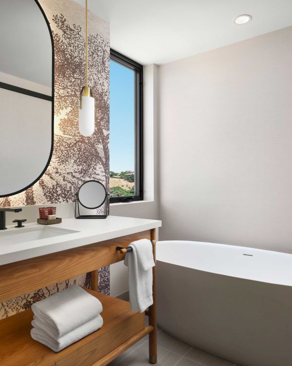 A modern bathroom features a freestanding tub, wooden vanity with a sink, towels, a round mirror, and a window with an outdoor view.