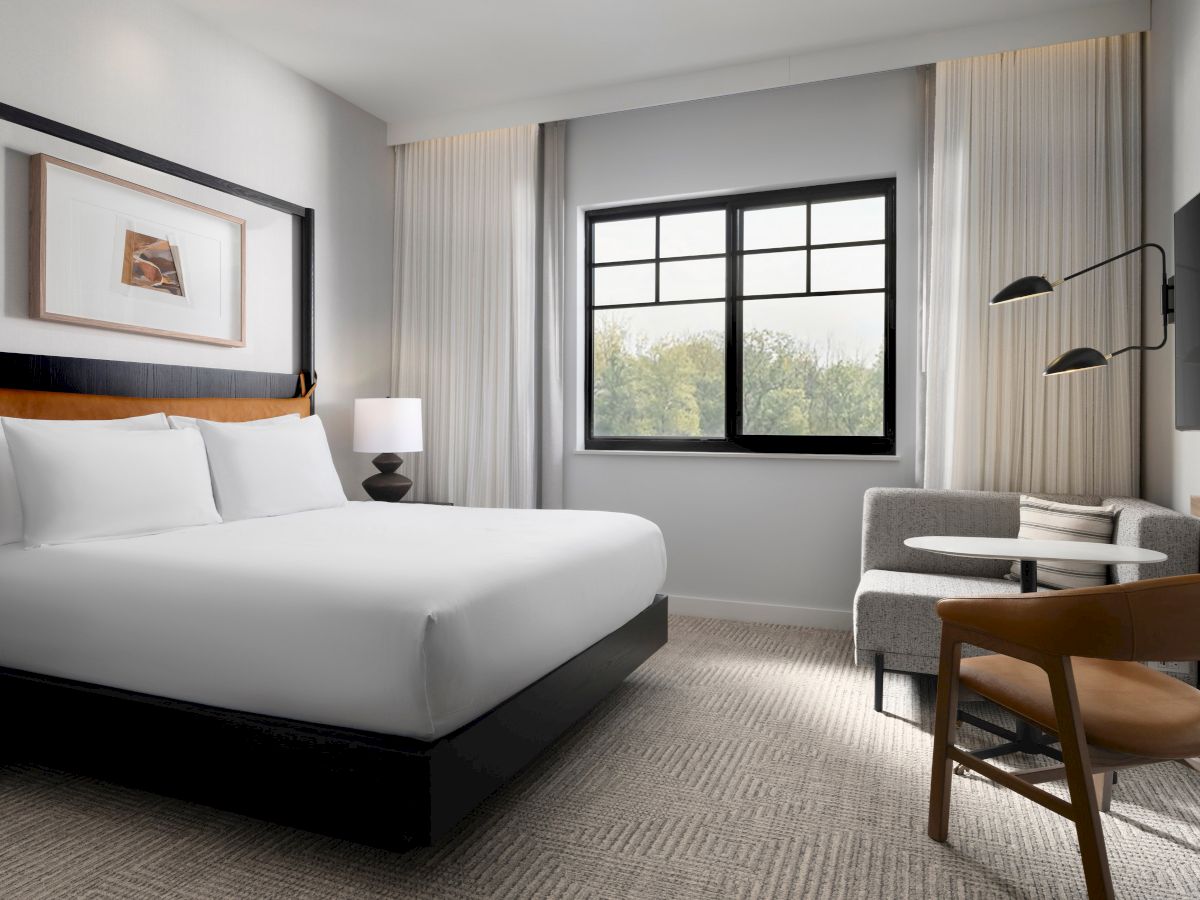 A modern, minimalist hotel room with a bed, artwork on the wall, a side chair, table, and a window with a view.