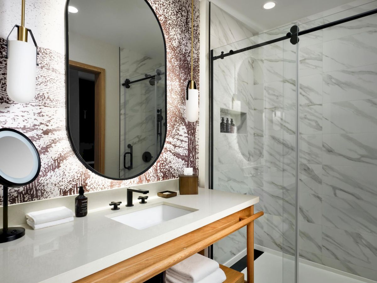 Modern bathroom with marble shower, oval mirror, vanity with black fixtures, and lighting. Towels and soap bottles are on the counter.
