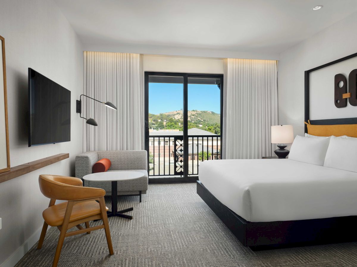A modern hotel room with a large bed, TV, armchair, and sofa, facing a window with a scenic view.